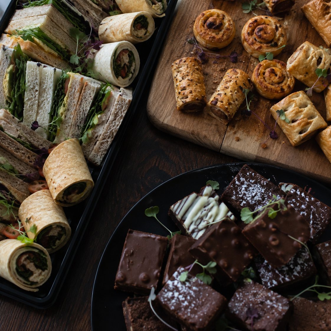 A catering spread on a dark table: a black tray of sandwiches and wraps, a wooden board of sausage rolls and savoury swirls, and a plate of brownies